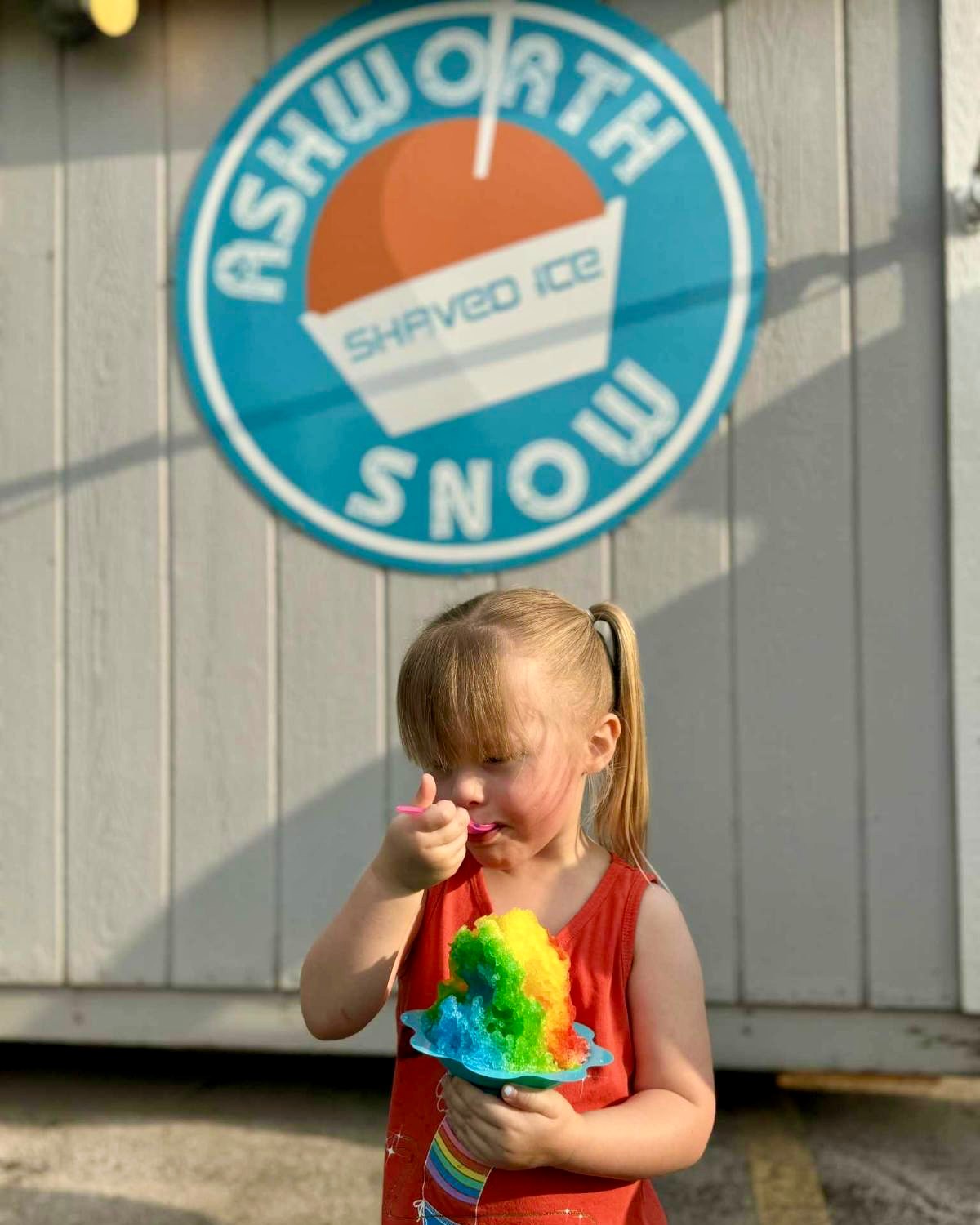 Little girl with rainbow shaved ice in front of the Ashworth Snow logo
